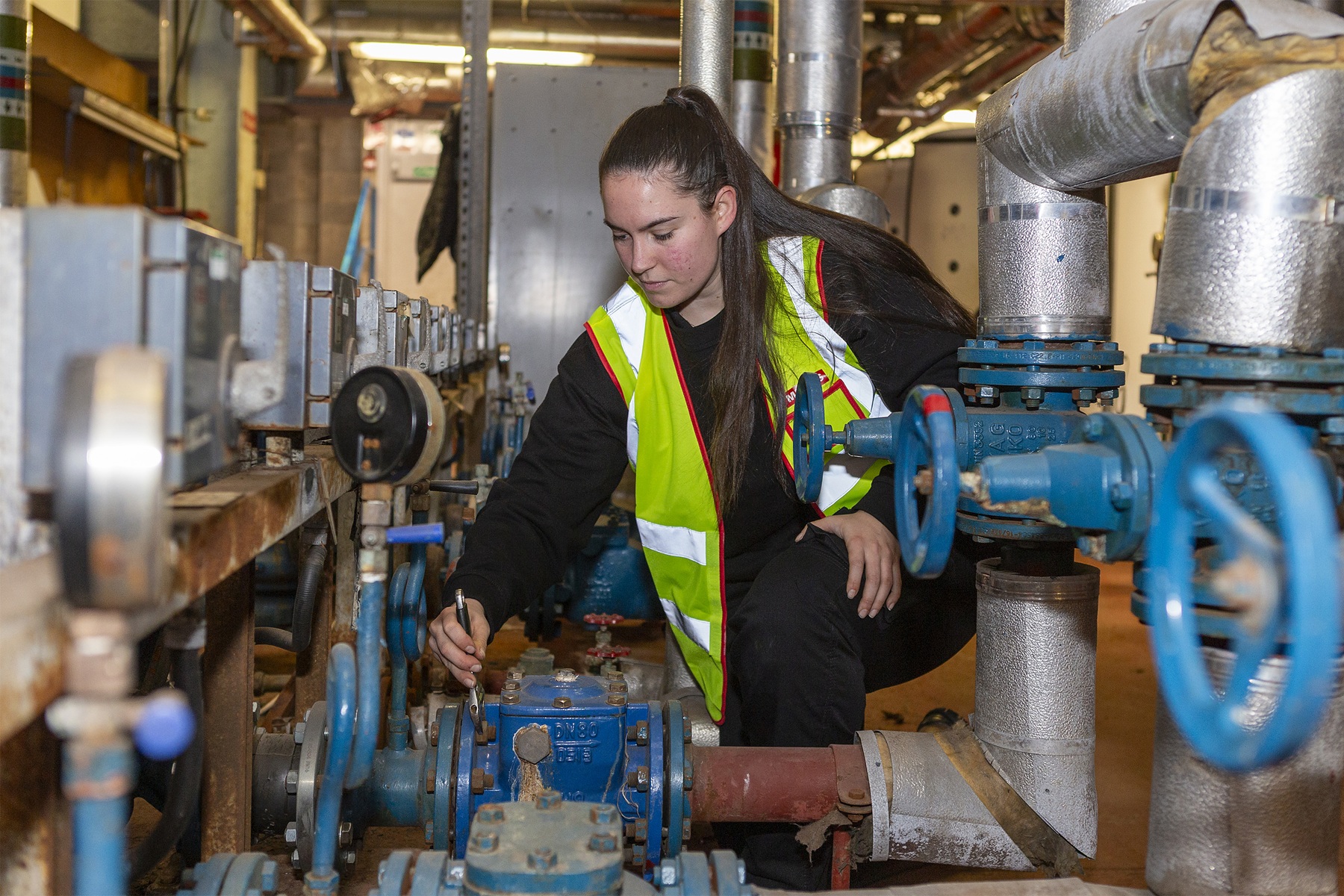 A person wearing a high‑visibility vest kneels in an industrial plant room, using a wrench to adjust a blue valve on a network of pipes and machinery.
