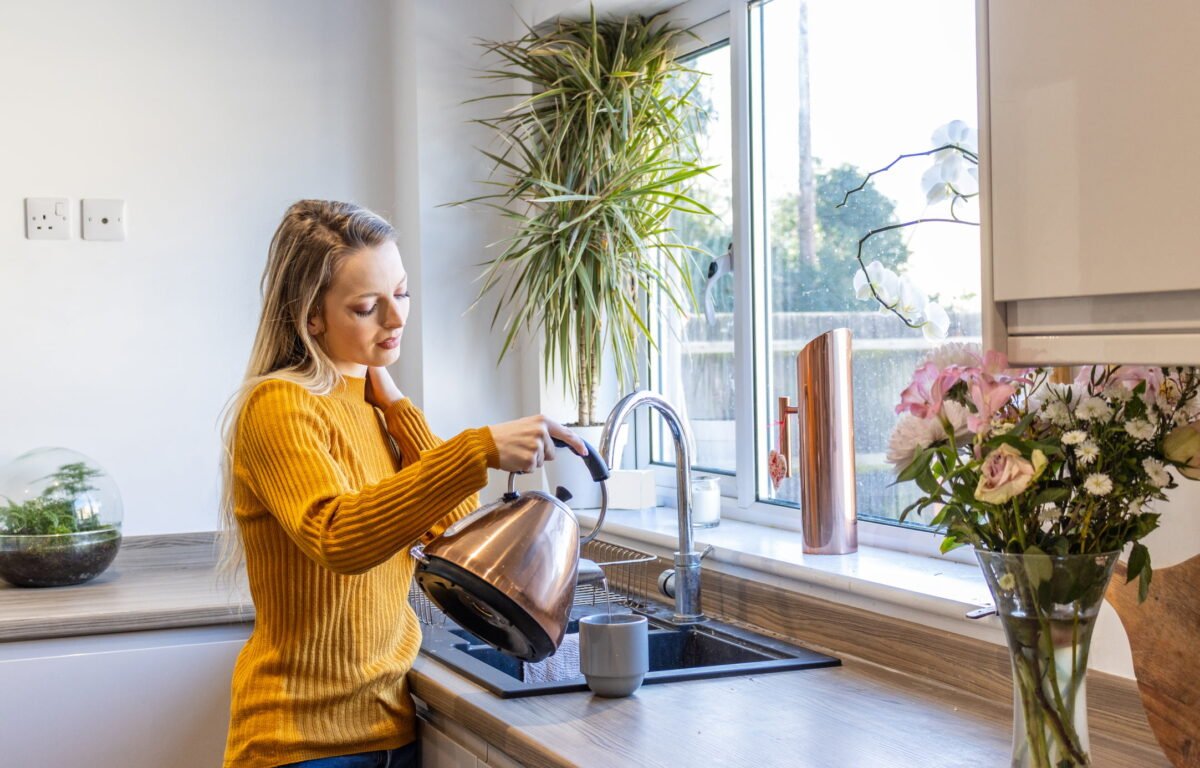 Blond young woman stands at the kitchen window and pours some water from a kettle into a mug