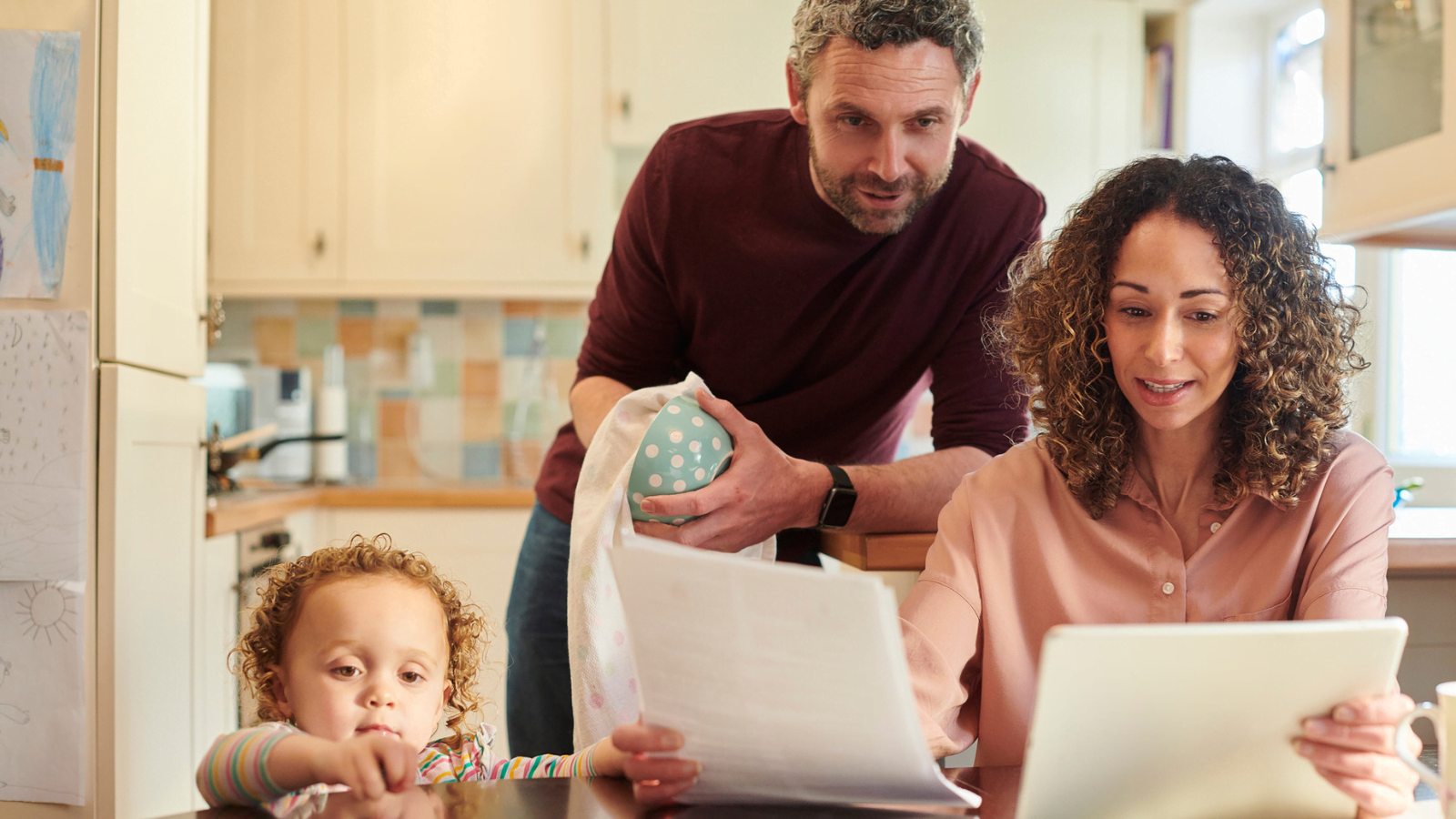 Couple with baby sat at kitchen table looking at paper bill and digital tablet