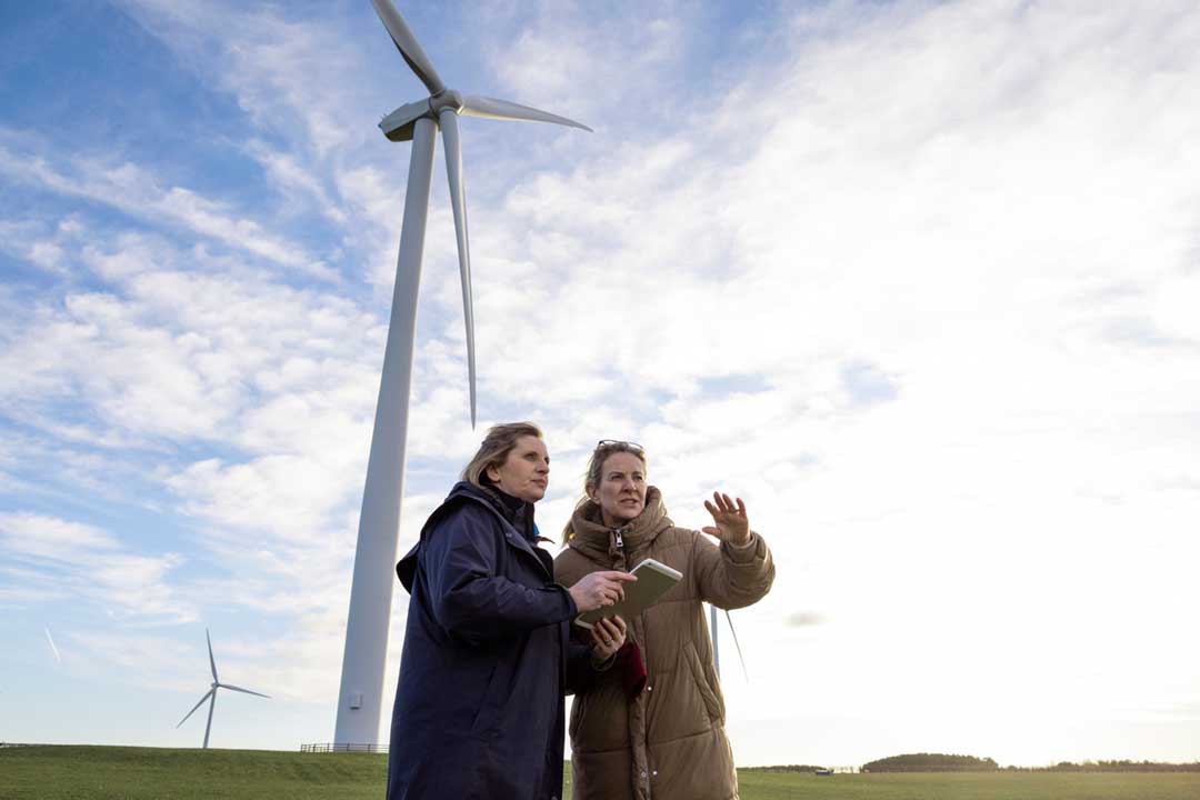Two women wearing jackets stood in front of a wind turbine.