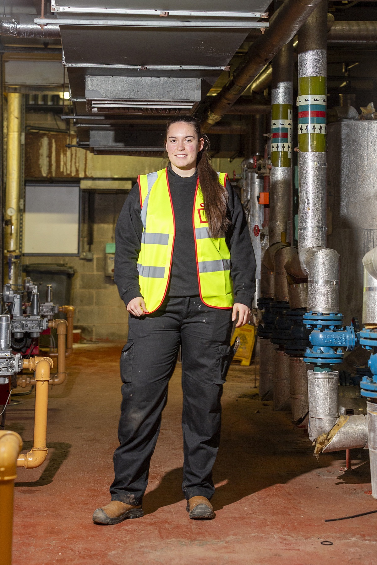 A person wearing a high‑visibility vest and work trousers stands in a plant room surrounded by large industrial pipes, valves, and ducting.