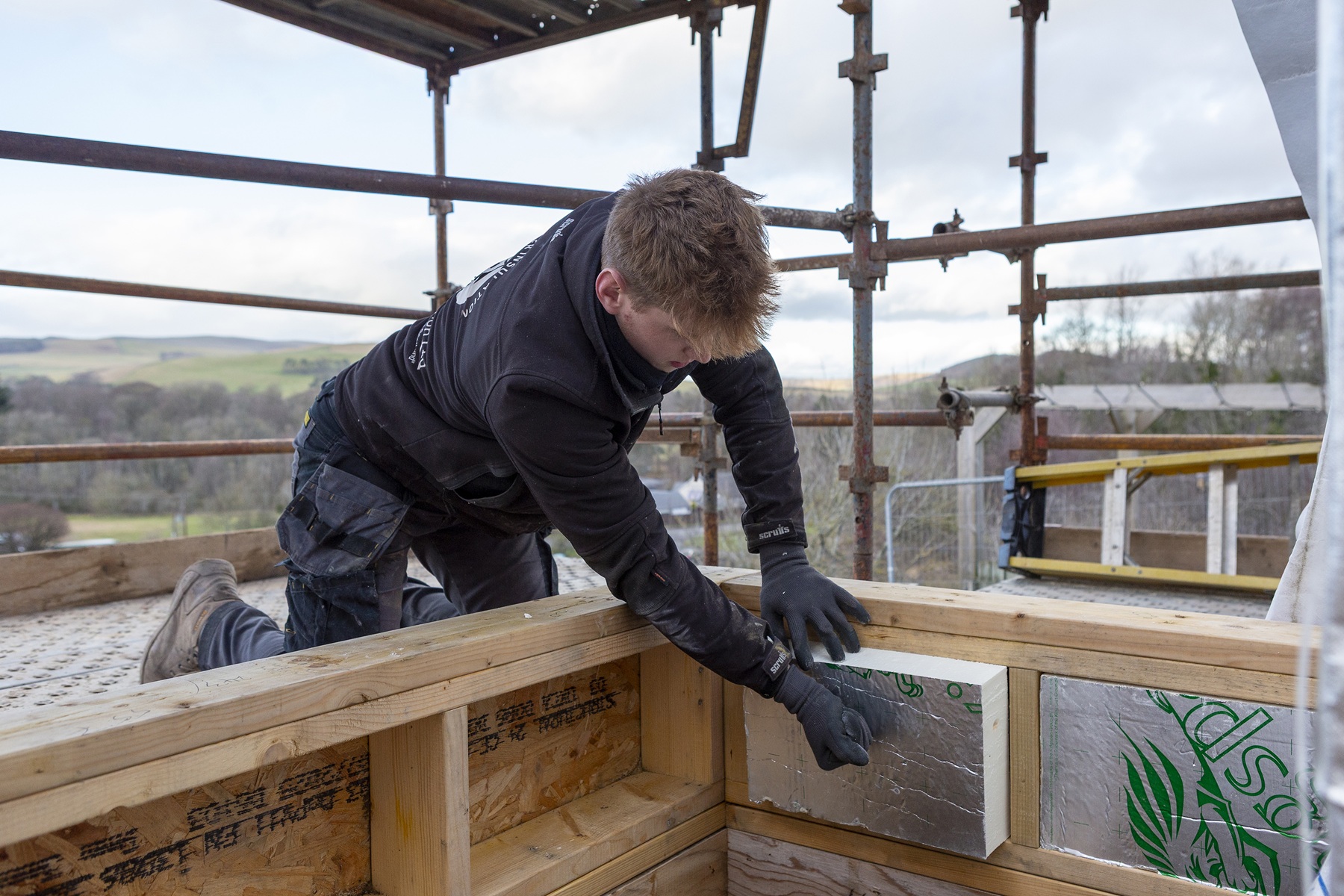 A worker kneels on a scaffold platform and fits a rigid insulation panel into a timber frame, with open countryside visible in the background.