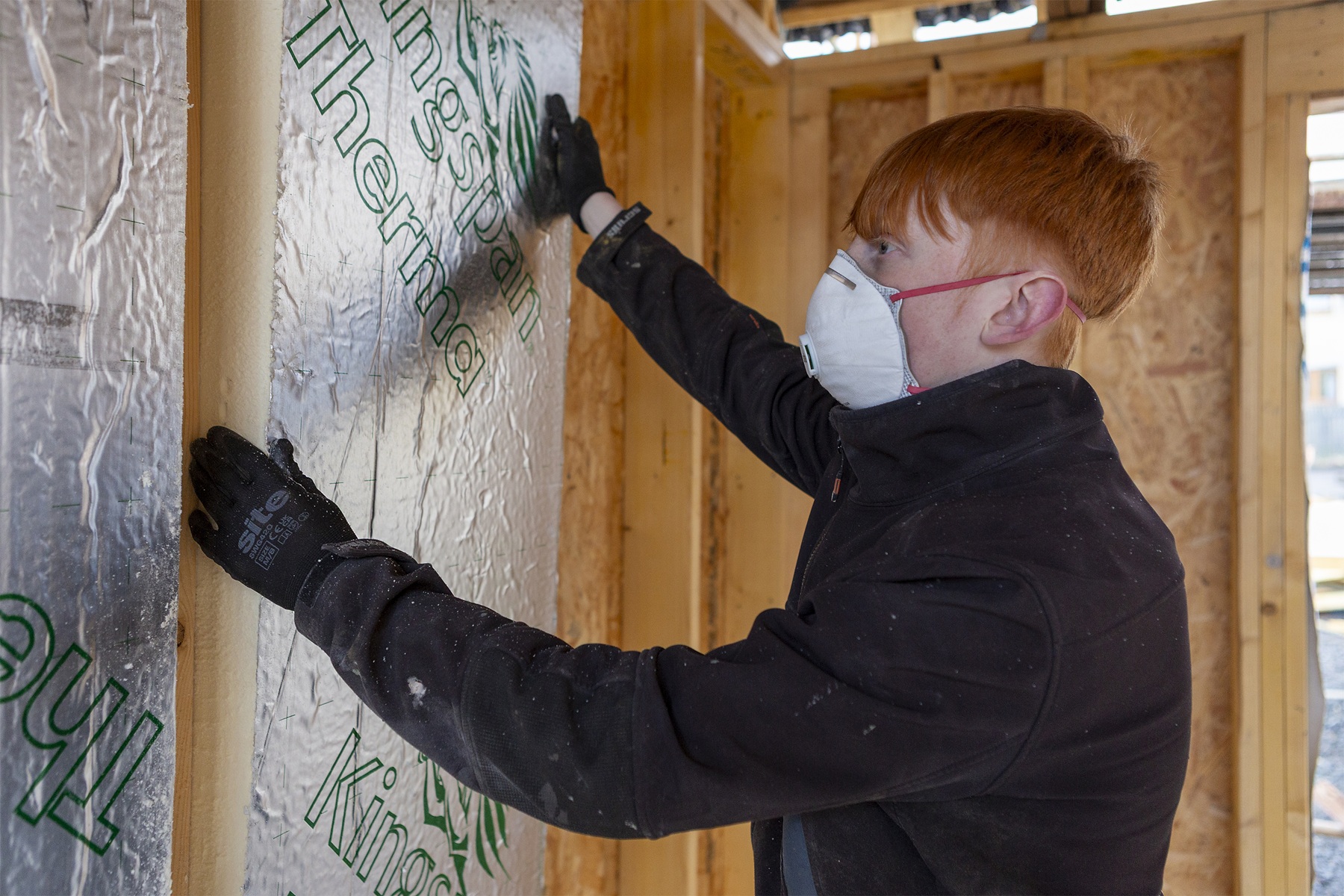 A worker wearing gloves and a dust mask fits a rigid insulation board into a timber wall frame inside a building under construction.