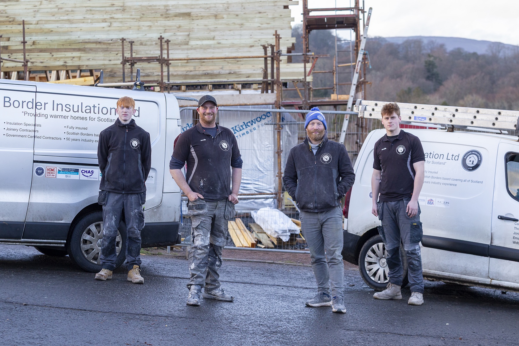 Four workers in construction clothing stand in front of two branded vans at a building site, with scaffolding and timber framing visible in the background.