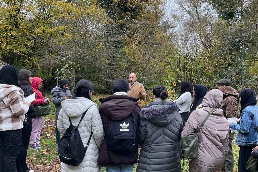 A group of people in coats ather around a central speaker in a woodland clearing on a fine day.