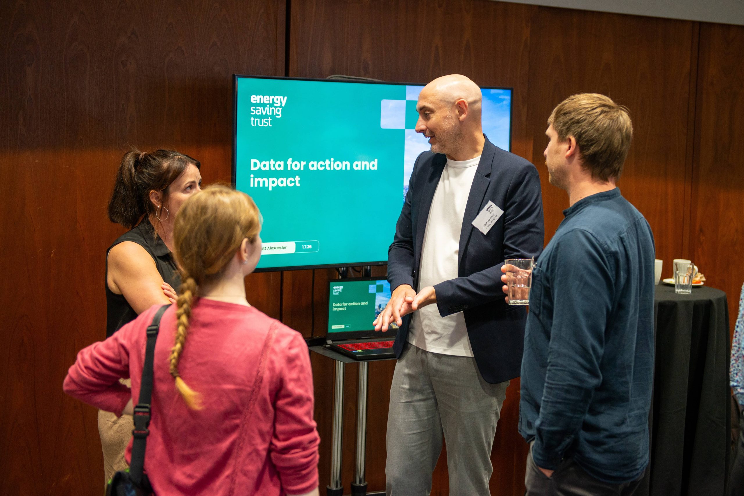 Matt Alexander from Energy Saving Trust speaking to three delegates from the Warm Homes Plan event, in front of a screen with a PowerPoint presentation slide with the Energy Saving Trust logo displaying the words: Data for action and impact.