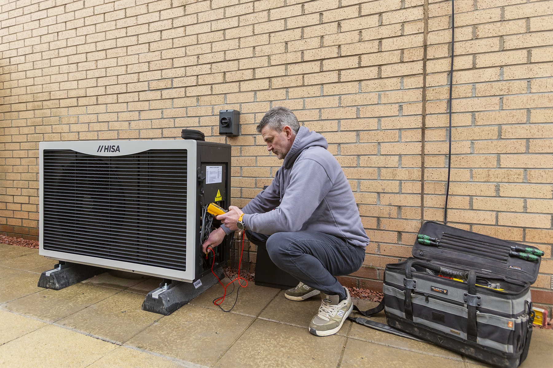 Person servicing an outdoor air source heat pump beside a building, with testing equipment and tools nearby.