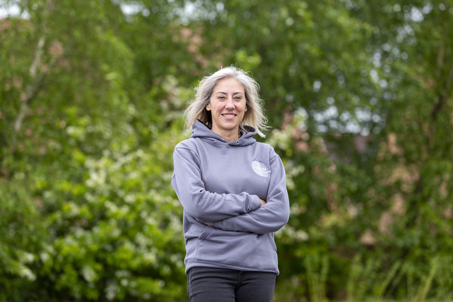 Person with shoulder-length hair stands outdoors with arms crossed, wearing a grey hoodie and dark trousers. Green trees and foliage form a blurred background.