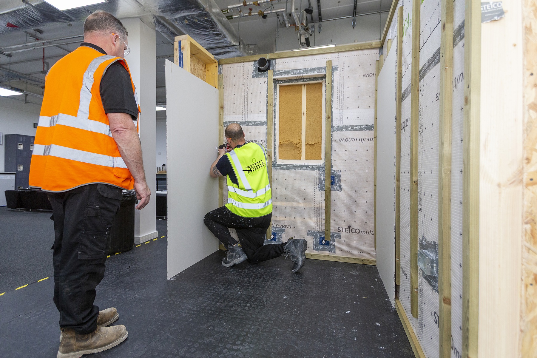 Two people installing plasterboard panels on a timber-framed training wall structure.