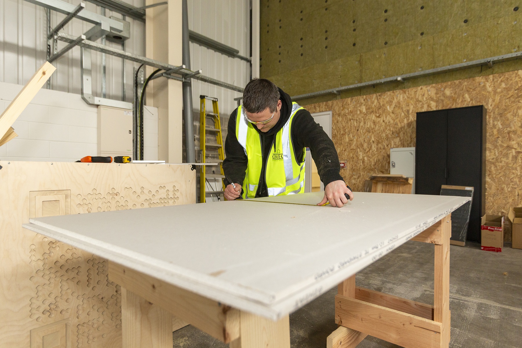 Person in a high-visibility vest measuring a plasterboard sheet on a workbench in a workshop.