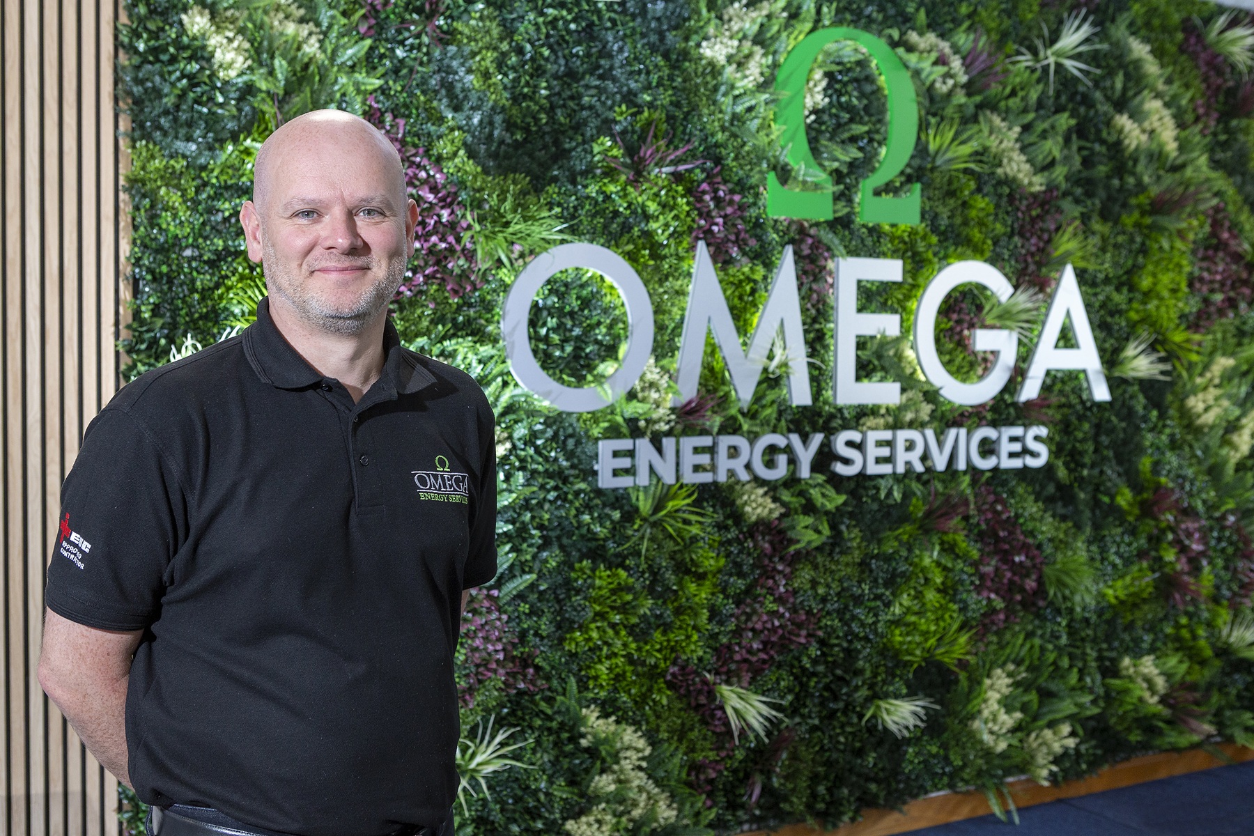 Person in an Omega Energy Services polo shirt standing in front of an Omega Energy Services branded green living wall.